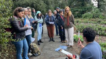 a photo of a man crouching down in front of a group of people holding a clipboard and a trowel