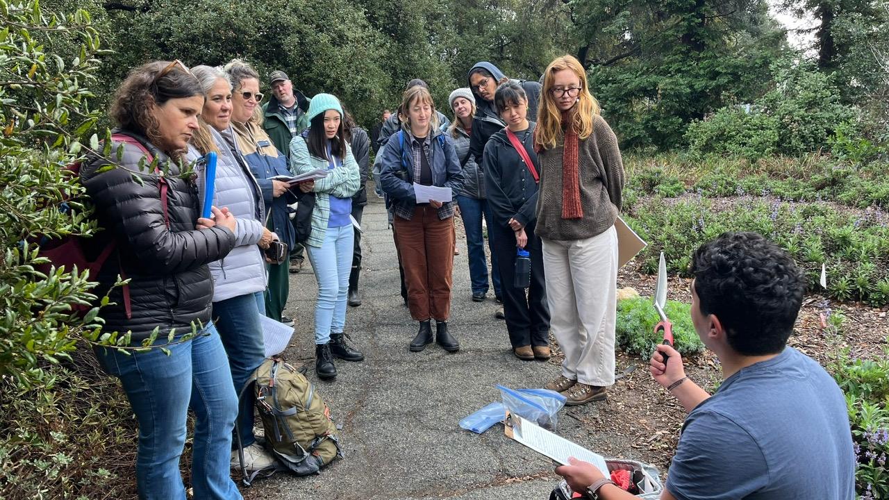 a photo of a man crouching down in front of a group of people holding a clipboard and a trowel