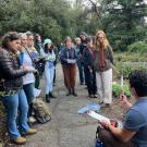a photo of a man crouching down in front of a group of people holding a clipboard and a trowel